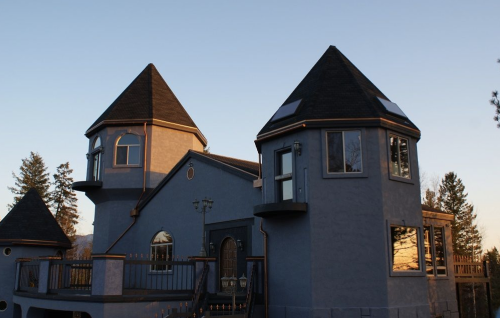 A unique two-towered house with a blue exterior, surrounded by trees and bathed in warm sunset light.