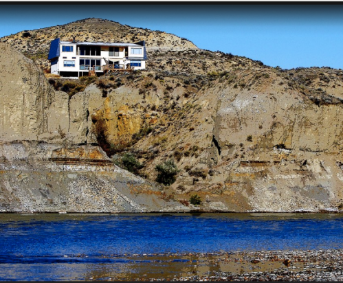 A modern house perched on a rocky cliff overlooking a calm blue river.