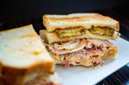 A close-up of a toasted sandwich filled with layers of turkey, ham, cheese, and pickles on a white plate.