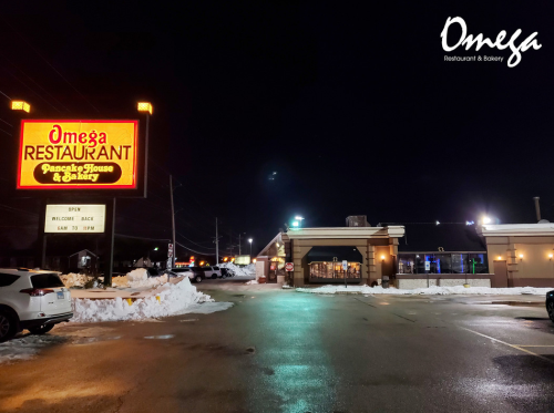 A brightly lit Omega Restaurant and Bakery at night, with snow on the ground and a welcoming sign.