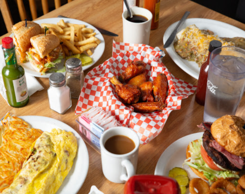 A table spread with sandwiches, fries, wings, an omelet, a burger, coffee, and drinks in a casual dining setting.