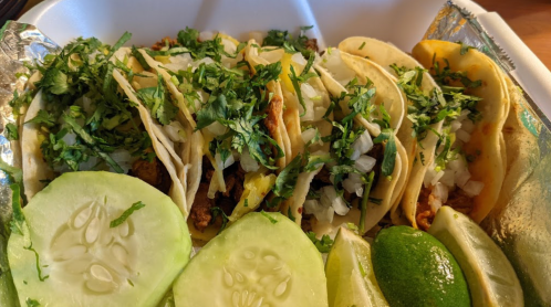 A takeout container with several tacos topped with cilantro and onions, alongside cucumber slices and lime wedges.