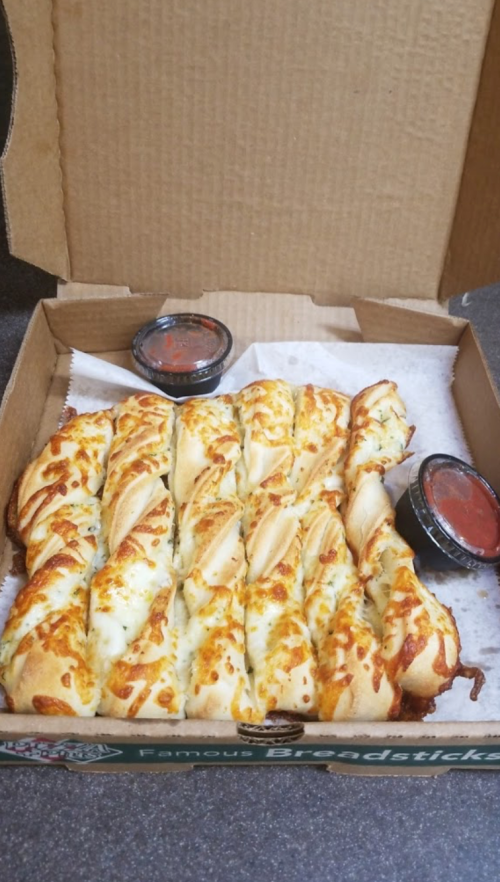 A pizza box containing cheesy breadsticks with marinara sauce on the side.
