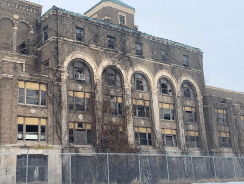 Abandoned brick building with arched windows, overgrown vines, and a chain-link fence in front.