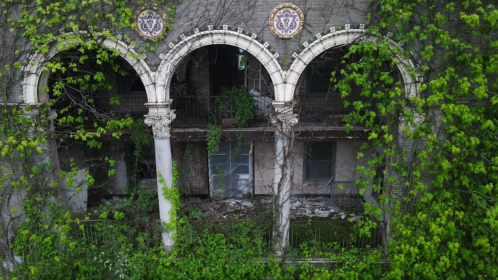 Abandoned building with arched columns, overgrown with vines and greenery, showcasing decay and nature's reclamation.