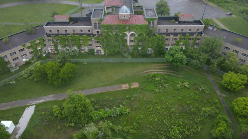 Aerial view of an abandoned building covered in greenery, surrounded by overgrown land and trees.