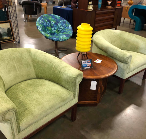 Two green upholstered chairs with wooden legs, a round wooden table, and a yellow lamp in a vintage furniture store.