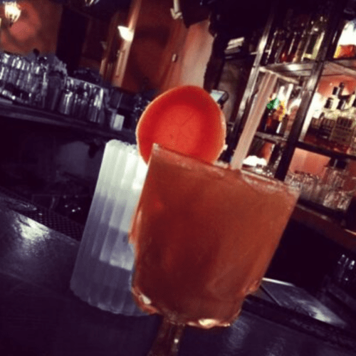 A close-up of a cocktail glass with a peach slice, set against a blurred bar background.