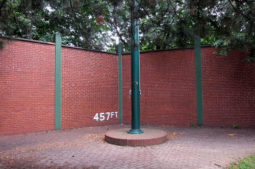 A brick wall with a green pole in the center, marked with "457FT," surrounded by trees.
