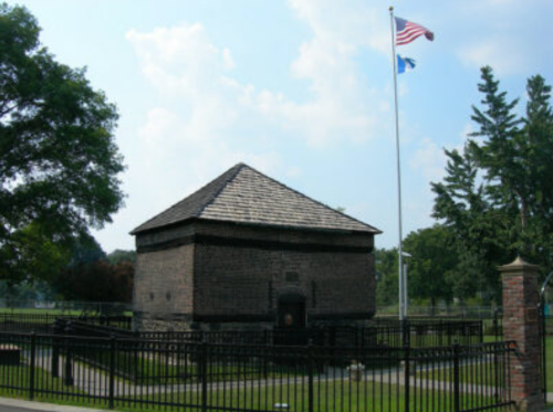 Historic stone building with a peaked roof, surrounded by a black fence, under a blue sky with flags flying.