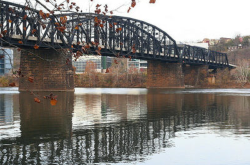 A steel bridge spans a calm river, reflecting its structure, with autumn leaves visible in the foreground.