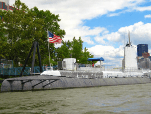 A submarine with an American flag docked near a waterfront, surrounded by trees and buildings under a cloudy sky.