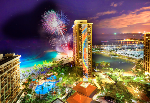 Colorful fireworks illuminate the night sky over a beachfront resort, with a pool and marina visible below.