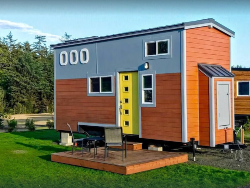 A colorful tiny house with orange and blue siding, a yellow door, and a small deck with chairs in a grassy area.