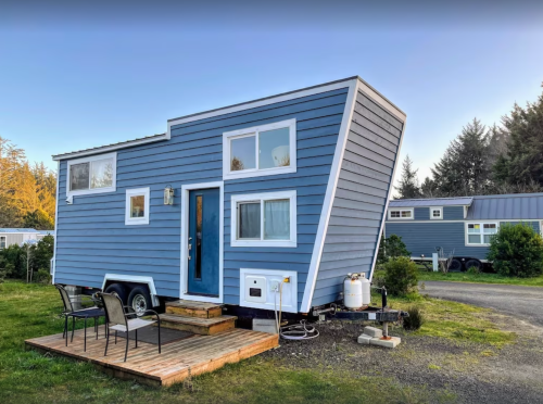 A blue tiny house on wheels with a unique angled design, set on a grassy area with a wooden deck and chairs.