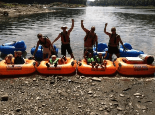 A group of people celebrating on the shore with inflatable rafts by a river, enjoying a sunny day outdoors.