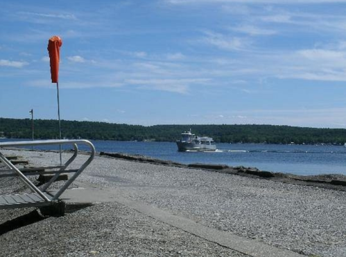 A ferry sails across a calm lake on a sunny day, with a sandy shore and a windsock in the foreground.