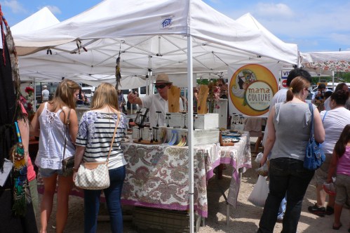 A bustling market scene with vendors under white tents, showcasing crafts and products to interested shoppers.