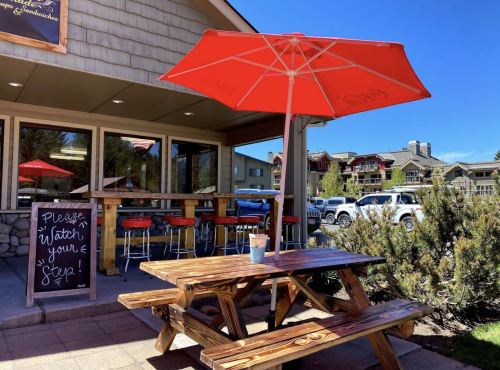 A wooden picnic table with an orange umbrella outside a café, featuring a sign that says "Please Watch Your Step."