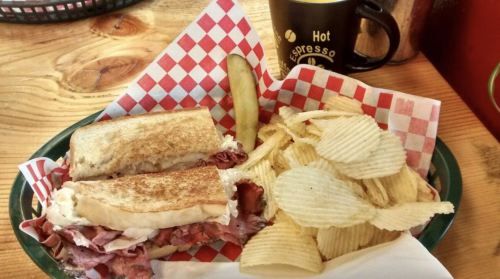 A plate with a toasted sandwich, pickles, and a side of potato chips, alongside a coffee cup.
