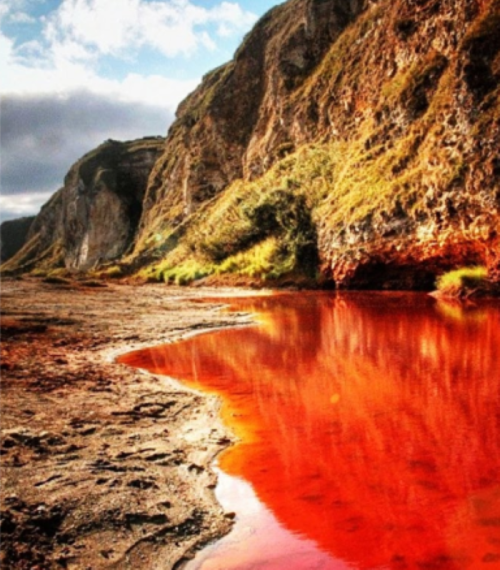 A scenic landscape featuring red-tinted water reflecting the surrounding cliffs and greenery under a cloudy sky.