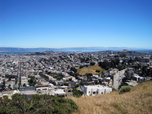 A panoramic view of a cityscape with hills, water, and clear blue skies in the background.