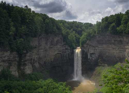 A majestic waterfall cascades down rocky cliffs, surrounded by lush green trees and a cloudy sky.