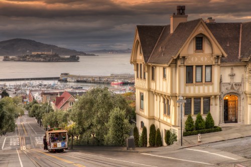 A scenic view of a steep street with a cable car, overlooking a harbor and Alcatraz Island under a cloudy sky.