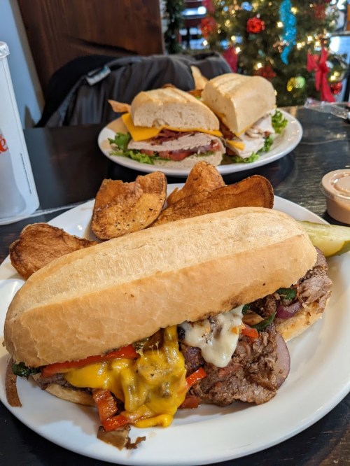 A hearty sandwich with melted cheese, peppers, and beef, served with crispy chips, with another sandwich in the background.