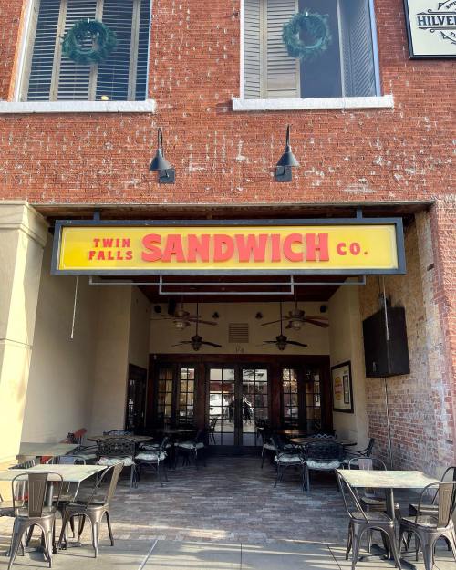Exterior of Twin Falls Sandwich Co. with a bright yellow sign, featuring outdoor seating and brick walls.