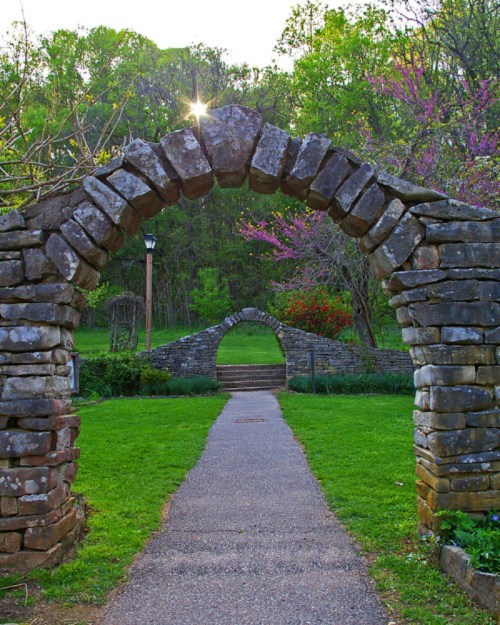 A stone archway frames a pathway leading to another arch, surrounded by lush greenery and blooming trees.