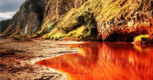 A scenic landscape featuring red-tinted water reflecting cliffs and greenery under a cloudy sky.