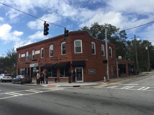 A brick building at an intersection with traffic lights, featuring shops and people outside on a cloudy day.