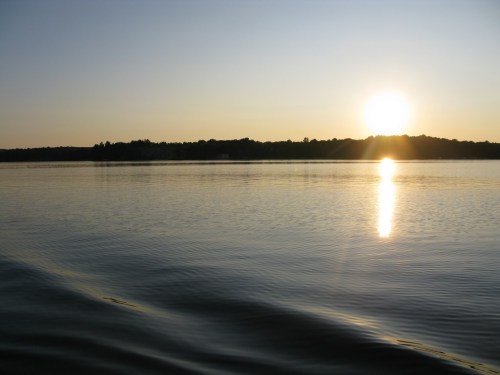 A serene sunset over a calm lake, with golden reflections on the water and silhouettes of trees in the background.