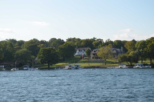A serene lake view with lush green trees, a large house on a hill, and small boats near the shore.