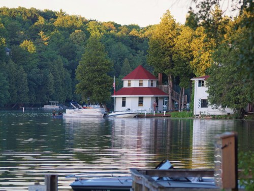 A serene lakeside scene featuring a house with a red roof, boats, and lush green trees reflecting in the water.