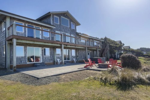 A coastal house with large windows, a patio, and red chairs around a fire pit, surrounded by grass and shrubs.