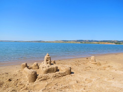 A sandcastle on a sandy beach by a calm lake, with clear blue skies and distant hills in the background.