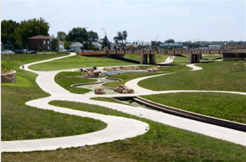 A winding pathway through a green park with a bridge in the background and rocks along the water's edge.