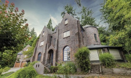 A unique brick house with a gothic design, surrounded by lush greenery and tall trees under a cloudy sky.