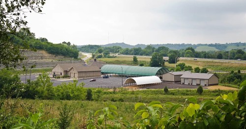A view of a rural facility with multiple buildings, surrounded by greenery and hills, near a highway.