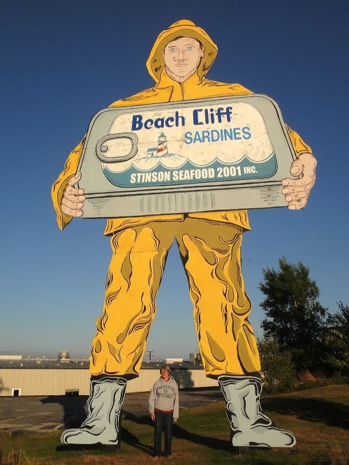 A person stands next to a giant, colorful sardine can sign featuring a fisherman in a yellow raincoat.