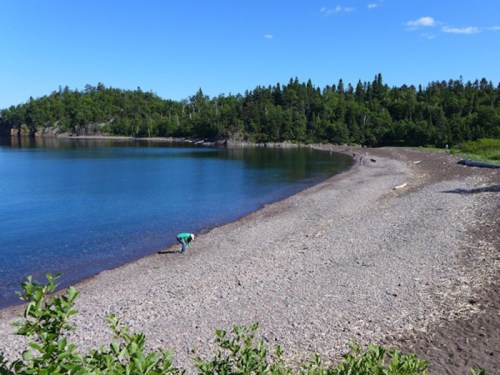 A serene beach with a pebbly shore, surrounded by lush green trees and a calm blue water, with a person exploring the shoreline.