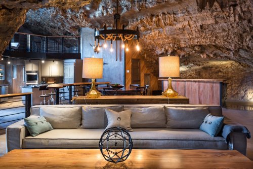 A cozy cave interior featuring a stylish sofa, wooden table, unique lighting, and a modern kitchen in the background.