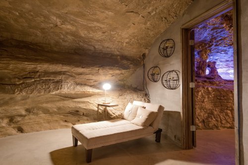 A cozy lounge area in a cave, featuring a chaise lounge, lamp, and decorative wall accents. Soft lighting enhances the ambiance.