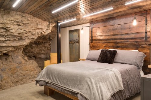 Cozy bedroom with a stone wall, wooden accents, and modern lighting, featuring a plush bed and a yellow armchair.