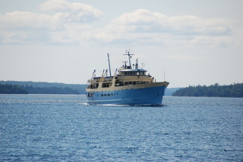 A blue and yellow ferry boat sails across a calm lake with trees in the background under a clear sky.