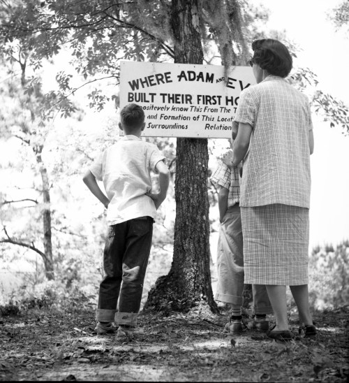 A woman and two children stand by a sign reading "Where Adam and Eve Built Their First Home" in a wooded area.