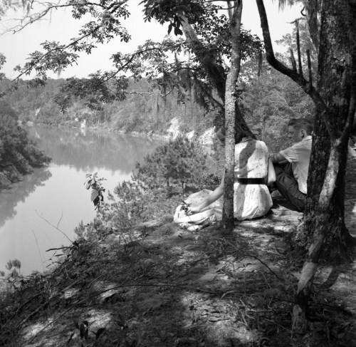 A black and white photo of a person sitting on a rock by a river, surrounded by trees and nature.