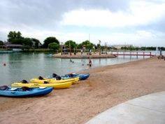 Colorful kayaks on a sandy beach by a calm waterway, with people swimming and a small island in the background.
